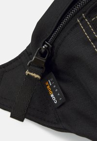 Melnas auduma soma ar rāvējslēdzēju, Cordura uzlīmi un teksturētu virsmu ar redzamām šuvēm un aksesuāriem papildu izturībai.