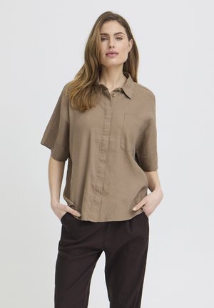 Kvinde med langt hår iført løs brun kortærmet skjorte med brystlomme og sorte bukser, hænderne i lommerne, stående mod en ensfarvet baggrund.