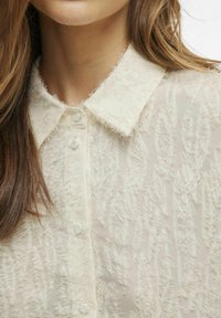 Blusa de crema claro con un tejido texturizado y acentos deshilachados, cuello puntiagudo y cierre de botones en la parte delantera.