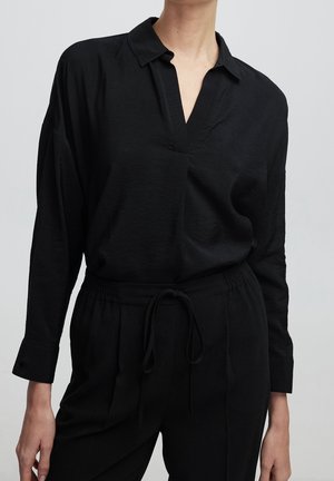 Femme portant un chemisier noir à manches longues avec un col en V et un pantalon noir assorti à taille élastiquée avec cordon.