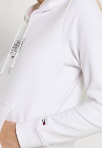 Bílá mikina s kapucí vyrobená z bavlněné směsi s potiskem loga a šňůrkami. Má boční kapsu a malou barevnou blokovou nášivku na manžetě.