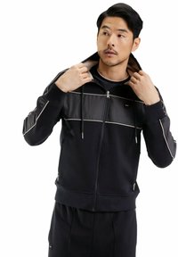 Homme ajustant la capuche d'une veste zippée noire avec des bordures beiges, portant un pantalon noir assorti, regardant sur le côté devant un fond blanc.