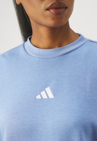 Ljusblå sweatshirt med rundad halsringning och mjuk textur, med en vit broderad Adidas-logotyp på framsidan.