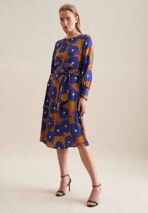 Bloemjurk met een mosterdachtergrond, voorzien van grote blauwe bloemen, witte accenten, lange mouwen en een taillebanddetail. Zwarte bandjeshakken.