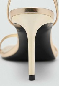 Gold strappy high-heel sandal with a pointed toe and a sleek black sole. The heel is metallic and slender, tapering to a small base.