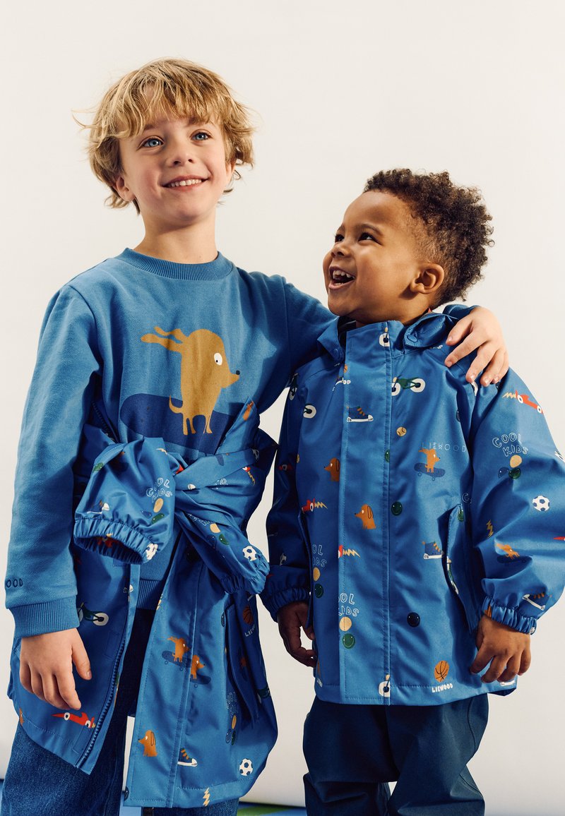 Deux enfants portent des vêtements bleus ; l'enfant plus âgé est vêtu d'un sweat-shirt avec un motif de chien, tandis que le plus jeune porte un imperméable à motifs colorés.