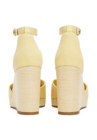 Geel canvas wedge schoenen met een gestructureerd beige plateau en een verstelbaar enkelbandje; de achteraanzicht benadrukt het verticale hakontwerp.