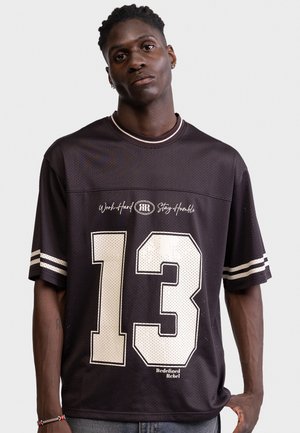 Ung mand iført sort sportstrøje med hvide striber, nummer 13 og teksten "Work Hard Stay Humble" samt "Redefined Rebel" synligt.