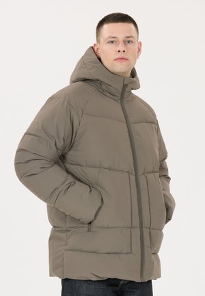 Mand iført en taupe hættetrøje med hættes, hænderne i lommerne, stående mod en lys baggrund.