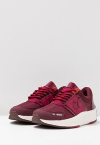 Bordowe buty sportowe z teksturowaną siateczką, zamszowymi akcentami i grubą białą podeszwą; zawierają pasujące sznurowadła i detal z logo.