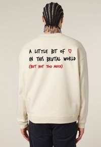 Felpa color crema con polsini e orlo a coste. La parte posteriore presenta un testo in stile scritto a mano in nero e rosso, che trasmette un messaggio.