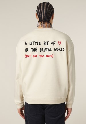 Crème-kleurige sweatshirt met ribgebreide manchetten en zoom. De achterkant heeft handgeschreven tekst in zwart en rood, die een boodschap overbrengt.