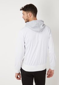 Chaqueta con capucha de tejido gris claro con una textura suave, que presenta una cinturilla y puños en blanco, mostrada desde atrás.
