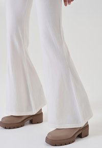 Pantaloni bianchi a palazzo con una texture liscia e orlo cucito, abbinati a stivali marroni con tacco alto e suola rigata.