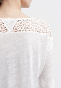 Bílý lehký top s texturovaným krajkovým zadním dílem, s jemným výstřihem do V a volným střihem. Měkký, prodyšný materiál.