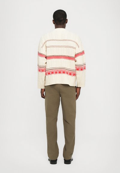 Jersey de punto crema con rayas rojas y marrones, que presenta un patrón texturizado. Combinado con pantalones verde olivo y zapatos negros.