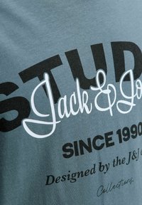 Kortærmet t-shirt i teal farvet bomuld med sort og hvid grafisk tekst: "STUD" og "Jack & Jones" samt en glat overflade.