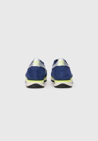 Dvojica modrých semišových tenísk Adidas so striebornými pätkami a neonovo žltými akcentmi, zobrazené zozadu na jednoduchom bielom pozadí.