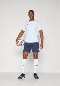 T-shirt de sport blanc avec des accents noirs, shorts navy, chaussettes blanches et chaussures de foot noires. Tenant un ballon de football, debout devant un fond gris.