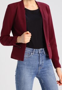 Blazer bordeaux à la coupe ajustée, manches longues, sans col, avec des accents de fermeture éclair latérale, assorti à un haut noir et un jean bleu.