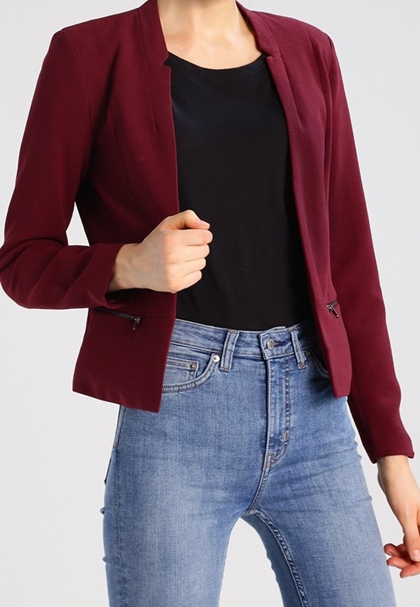 Blazer bordeaux à la coupe ajustée, manches longues, sans col, avec des accents de fermeture éclair latérale, assorti à un haut noir et un jean bleu.
