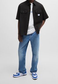 Zwart shirt met korte mouwen en twee voorzakken, wit T-shirt eronder, blauwe spijkerbroek en blauw-witte sneakers.