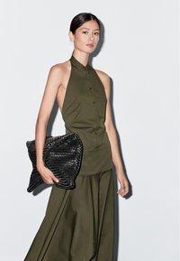 Olivengrøn halterneck-top med knaplukning foran, parret med en svajende nederdel og en stor sort struktureret clutch-taske. Glat stoffinish.
