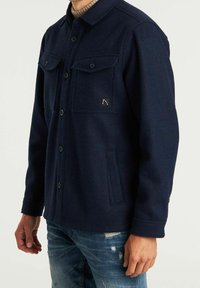 Mann trägt eine marineblaue, geknöpfte Wolljacke mit Brust- und Seitentaschen über einem beigen Rollkragenpullover und zerrissenen blauen Jeans.