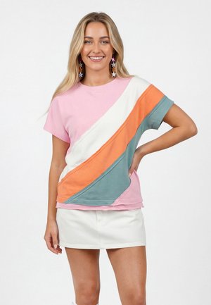 Uśmiechnięta kobieta o długich blond włosach, nosząca kolczyki w kształcie gwiazdek, różowy T-shirt z białymi, pomarańczowymi i niebieskimi ukośnymi paskami oraz białą spódnicę.