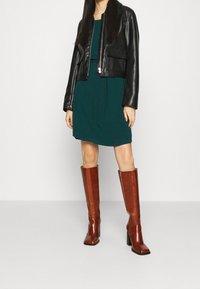 Czarna skórzana kurtka typu cropped, turkusowa sukienka z wiązaniem i brązowe, wysokie do kolan, botki na słupku. Gładkie tekstury, nowoczesny design.