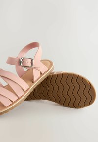 Sandali in pelle rosa chiaro con molte cinghie incrociate, fibbia regolabile e una suola in gomma marrone testurizzata con motivo a onde.