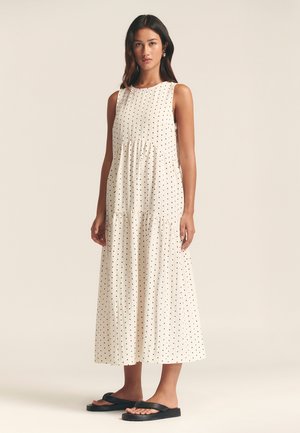 Sleeveless white dress with black polka dots, tiered design, textured fabric, and a relaxed fit, paired with black flat sandals.