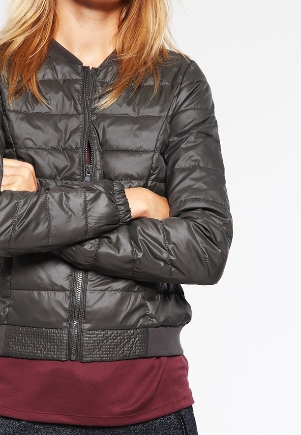 Femme les bras croisés, portant une veste matelassée noire zippée sur un t-shirt bordeaux, devant un fond uni.