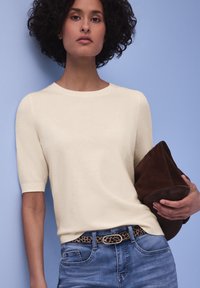 Femme aux cheveux bouclés portant un haut crème à manches courtes, un jean bleu avec une ceinture à imprimé léopard, tenant une grande pochette en daim marron.