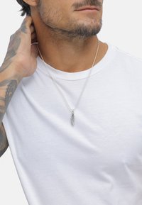 Sølvfjer pendul hænger fra en tynd kæde, der bæres over en ensfarvet hvid t-shirt. Huden har en let tan, med synlige tatoveringer på armen.