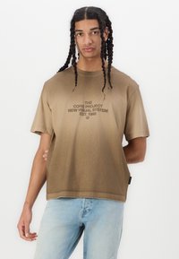 Brun gradient t-shirt med korte ærmer, der har trykt tekst i mørkebrun. Blødt stof, afslappet pasform og synlige syninger.