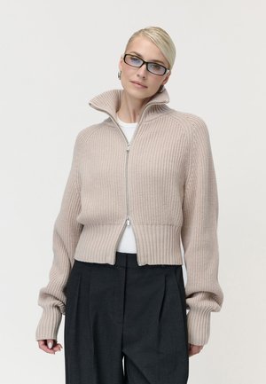 Kvinde med blondt hår og briller iført en beige strikket trøje med lynlås og mørke højtaljede bukser mod en lys baggrund.