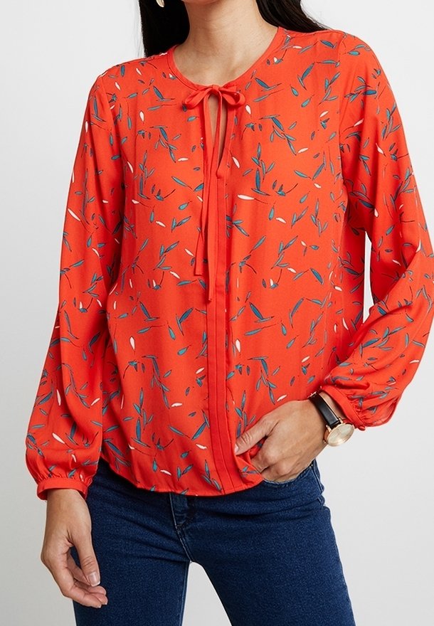 Blouse rouge à manches longues avec un motif floral bleu et blanc. Comprend un détail de nouage au niveau du cou et des poignets légèrement froncés.