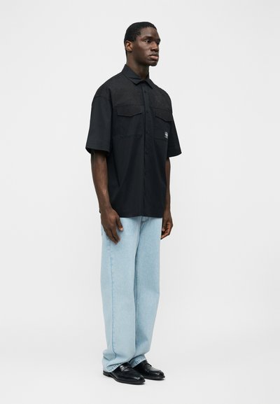 Muž stojící, oblečený v černé košili s krátkým rukávem na knoflíky, světlé modré široké džínsy a černé společenské boty, na jednobarevném pozadí.