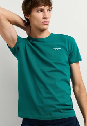 Joven con cabello corto y castaño que lleva una camiseta azul verdosa de Pepe Jeans, levantando su brazo izquierdo detrás de la cabeza, mirando hacia la derecha.