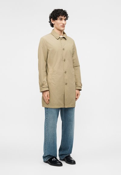 Άνδρας με σγουρά μαλλιά που φοράει μπεζ παλτό με κουμπιά, μπλε τζιν και μαύρα παπούτσια, στέκεται μπροστά σε ένα απλό ανοιχτόχρωμο φόντο.