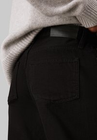 Crne traperice glatke teksture, s dva stražnja džepa i crnom kožnom etiketom s natpisom "Calvin Klein" na struku.
