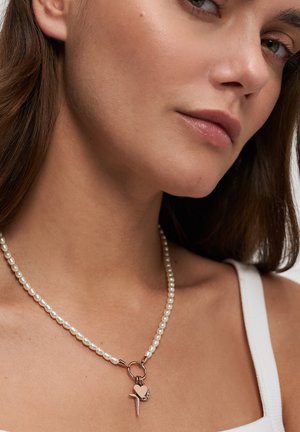 Nahaufnahme einer Frau, die eine weiße Perlenkette mit Anhängern in Form von Herz, Kreuz und Halbmond aus Gold trägt, kombiniert mit einem weißen Tanktop.