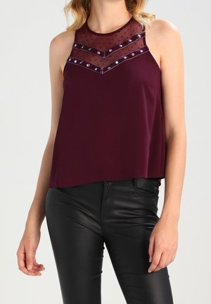 Ärmelloses dunkelviolettes Oberteil mit transparentem Tupfen-Einsatz und Verzierungen, kombiniert mit schwarzen Kunstlederhosen bei einer stehenden Person.