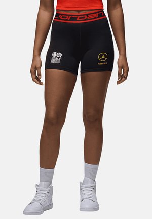 Athlet trägt schwarze Jordan-Shorts mit World Streetball-Logo und weiße High-Top-Sneaker mit Socken auf einfachem Hintergrund.