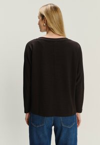 Blondynka w złotych kolczykach, ubrana w luźną, czarną bluzkę z długim rękawem i niebieskie dżinsy, odwrócona plecami w stosunku do jasnego tła.