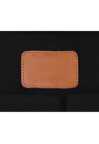 Toppa rettangolare in pelle color cuoio con cuciture visibili su sfondo in tessuto nero, centrata e leggermente rialzata.
