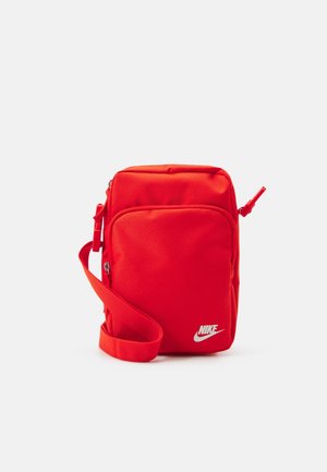 Kompaktowa czerwona torba na ramię Nike z regulowanym paskiem, przednią kieszenią na zamek i białym logo w prawym dolnym rogu.