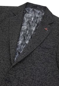 Blazer nero con tessuto testurizzato, caratterizzato da piccole macchie bianche. All'interno ha una fodera nera con un motivo a foglia e un piccolo logo rosso.