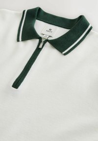 White polo shirt with a green collar featuring two white stripes, zipper closure, and tag labeled "NEXT" in size M. Textured fabric.